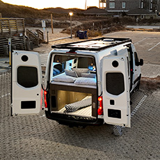 Fourgon aménagé stationné près d’une plage with portes arrière ouvertes, pillows on bed, and step laid out on sand